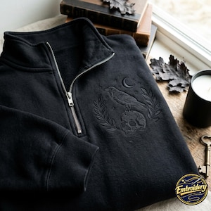 May include: A black quarter-zip sweatshirt featuring a tonal embroidered design on the chest. The embroidery depicts a raven perched on a human skull, surrounded by laurel branches, a crescent moon, and small stars. The zipper is silver-toned metal.