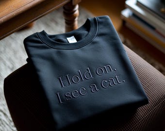 Può includere: Felpa nera con la frase ricamata "Hold on, I see a cat." La felpa di colore scuro è piegata e appoggiata su una sedia marrone a fantasia. È visibile lo scollo tondo.