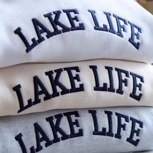 May include: A stack of three sweatshirts in white, beige, and gray. Each sweatshirt has the words "LAKE LIFE" embroidered in navy blue across the chest in a curved, block letter font. The sweatshirts are folded neatly.