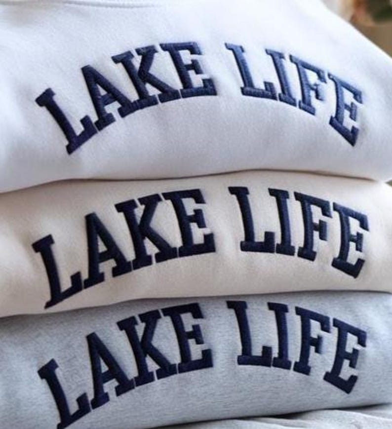 May include: A stack of three sweatshirts in white, beige, and gray. Each sweatshirt has the words "LAKE LIFE" embroidered in navy blue across the chest in a curved, block letter font. The sweatshirts are folded neatly.