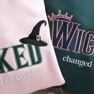 Puede incluir: Dos sudaderas, una rosa y otra verde azulado, bordadas con la palabra "WICKED" en una fuente estilizada. Cada sudadera presenta un sombrero de bruja y la frase "changed for good". Una corona est&aacute; bordada encima de la palabra "WICKED".