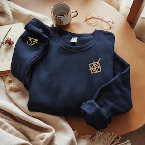 May include: A navy blue sweatshirt with gold embroidered details. The left sleeve features the number "33" in gold, while the chest displays a geometric design. The sweatshirt is folded on a wooden table, with a coffee cup and glasses in the background.