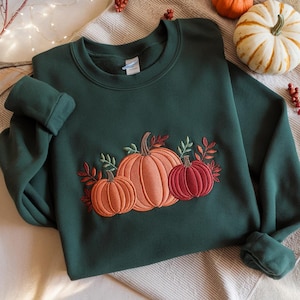 Peut inclure: Un sweat-shirt vert foncé avec une broderie de trois citrouilles aux tons orange et rouge, avec des feuilles vertes et orange. Les citrouilles sont disposées en rangée, la plus grande au centre.