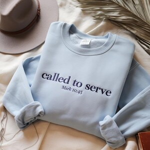 May include: A pale blue sweatshirt featuring the embroidered phrase "called to serve" and "Mark 10:45" in navy. The sleeves are rolled up. A grey hat and a book are visible in the background.