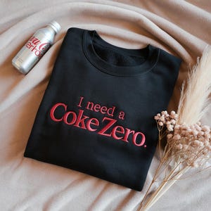 May include: Black crewneck sweatshirt with the embroidered phrase "I need a Coke Zero" in red. A silver Coca-Cola Zero aluminum bottle is visible on the left. The sweatshirt is displayed on a neutral-toned surface with dried flowers.