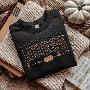 May include: Black sweatshirt with the word "NURSE" in a tan, outlined font. The design includes bats, a pumpkin, a candle, and a heartbeat line. The sweatshirt is displayed on a beige blanket with a white pumpkin and books.
