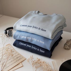 Könnte beinhalten: Ein Stapel von drei blauen Sweatshirts in Hellblau, Mittelblau und Marineblau. Auf jedem Sweatshirt steht der Text "Custom your lyrics in here" in Weiß. Die Sweatshirts liegen auf einer weißen Oberfläche.