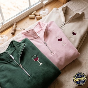 Puede incluir: Tres sudaderas con cremallera de un cuarto en verde oscuro, rosa claro y beige. Cada sudadera presenta un bordado de una copa de vino llena de vino tinto en el pecho izquierdo. Las prendas tienen cremalleras de metal plateado en el cuello.