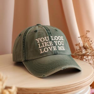 May include: An olive green baseball cap with the embroidered text "YOU LOOK LIKE YOU LOVE ME" in white. The cap has a curved brim and a distressed finish. A stylish accessory for everyday wear.