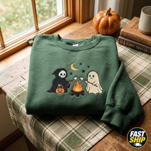 Op de afbeelding: Een bosgroene sweater met ronde hals en een geborduurd ontwerp van een Magere Hein met een pompoen en een spookje dat marshmallows roostert boven een kampvuur, met daarboven een gele maansikkel en enkele kleine zwarte sterren.