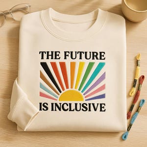 Puede incluir: Sudadera de color crema con el texto "THE FUTURE IS INCLUSIVE" sobre un diseño de rayos de sol colorido. El diseño de rayos de sol presenta un arcoíris de colores. La sudadera está doblada sobre una superficie de madera.
