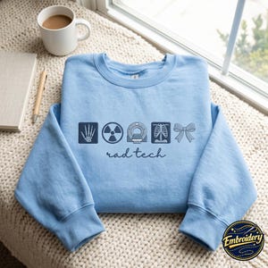 Puede incluir: Una sudadera azul claro de cuello redondo con bordado azul oscuro en el pecho. El diseño incluye iconos de un esqueleto de mano, un símbolo de radiación, un escáner CT, una caja torácica y un lazo, con el texto rad tech escrito debajo.