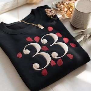 May include: Black sweatshirt with embroidered design. The design features the number "33" in a cream color, surrounded by red petal-shaped accents. A gold necklace with a pendant rests on the sweatshirt.