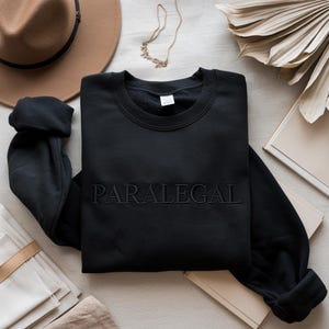 Puede incluir: Sudadera negra de cuello redondo con la palabra "PARALEGAL" bordada en el pecho. La sudadera se muestra sobre una superficie neutra con un sombrero marrón, un collar dorado y elementos decorativos.