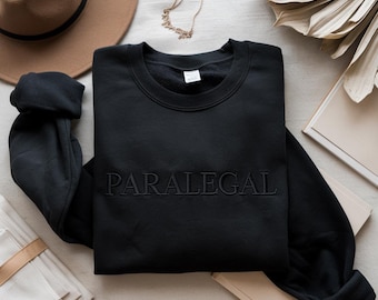 May include: Black crewneck sweatshirt with the word "PARALEGAL" embroidered across the chest. The sweatshirt is displayed on a neutral surface with a brown hat, gold necklace, and decorative elements.