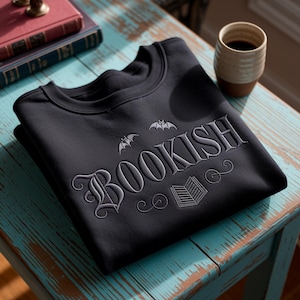 K&ouml;nnte beinhalten: Schwarzes Sweatshirt mit Rundhalsausschnitt und dem Wort "BOOKISH" in Grau gestickt, zusammen mit Fledermaus- und Buchgrafiken. Das Sweatshirt liegt gefaltet auf einem blau-verwitterten Tisch, mit einer Kaffeetasse und B&uuml;chern im Hintergrund.