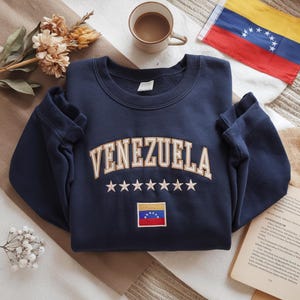 Op de afbeelding: Marineblauwe sweatshirt met het woord "VENEZUELA" in gouden, gebogen letters. Daaronder staan zes witte sterren en een kleine Venezolaanse vlag. De sweatshirt is op een patroonoppervlak gevouwen, met een kopje koffie en een Venezolaanse vlag op de achtergrond.