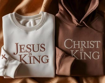 May include: A white crewneck sweatshirt with brown embroidered text reading JESUS IS KING and a brown hoodie with white embroidered text reading CHRIST IS KING. Both garments feature a small embroidered cross on the sleeve cuff.