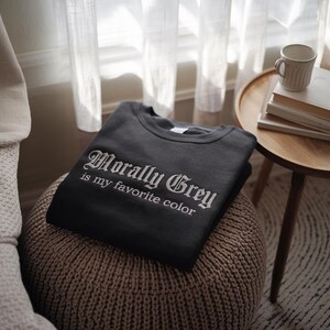 以下が含まれることがあります： 白いゴシック体で「Morally Grey is my favorite color」と書かれた、濃いグレーのスウェットシャツ。スウェットシャツは折りたたまれ、織りのオットマンの上に置かれています。マグカップと本が置かれた小さなサイドテーブルが背景にあります。
