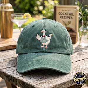Può includere: Un cappellino da baseball verde bosco con il ricamo di un'anatra bianca che indossa un gilet verde e un papillon bordeaux. L'anatra tiene uno shaker per cocktail argentato in un'ala e un bicchiere con una fetta di limone nell'altra ala.