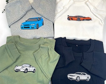 May include: Four sweatshirts with different car embroidery designs. The sweatshirts are in grey, white, green, and black. The car designs are a blue sports car, an orange sports car, a white sports car, and a white saloon.