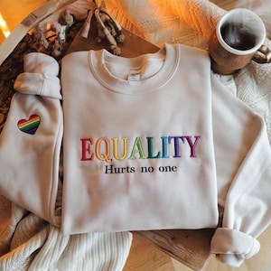 May include: A light beige sweatshirt with the word "EQUALITY" embroidered in rainbow colors. The text "Hurts no one" is embroidered below the word "EQUALITY" in black thread. A rainbow heart is embroidered on the left sleeve.
