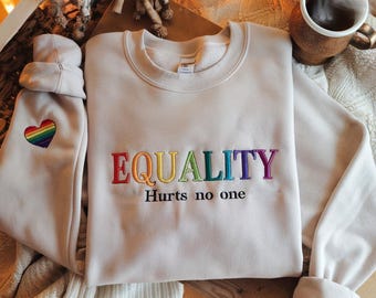 Può includere: Una felpa beige chiaro con la parola "EQUALITY" ricamata in colori arcobaleno. Il testo "Hurts no one" è ricamato sotto la parola "EQUALITY" in filo nero. Un cuore arcobaleno è ricamato sulla manica sinistra.