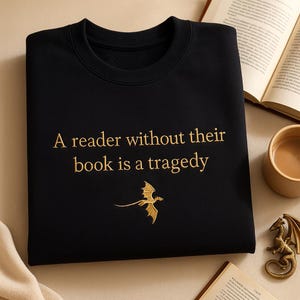 Op de afbeelding: Zwart sweatshirt met goud geborduurde tekst: "Een lezer zonder zijn boek is een tragedie." Een gouden draakafbeelding staat onder de tekst. Het sweatshirt ligt op een neutrale ondergrond met een boek en een kopje koffie.