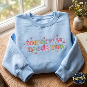 Puede incluir: Una sudadera de cuello redondo en color azul claro con el texto bordado "tomorrow needs you" en letras coloridas con degradado. El diseño está acentuado con pequeñas formas de estrellas bordadas multicolor que rodean las palabras.