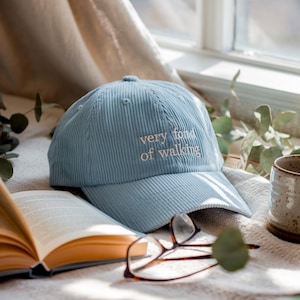 Könnte beinhalten: Hellblaue Cord-Baseballkappe mit dem gestickten Text "very fond of walking". Die Kappe liegt auf einer weichen Oberfläche neben einem offenen Buch und einer Brille. Eine Keramiktasse ist ebenfalls zu sehen.