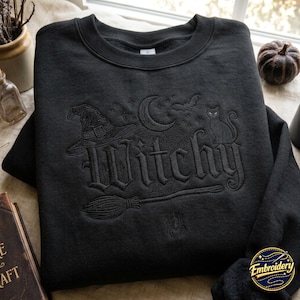 Könnte beinhalten: Ein schwarzes Sweatshirt mit Rundhalsausschnitt und einer tonalen schwarzen Stickerei des Wortes Witchy in gotischer Schrift. Das Design zeigt einen Hexenhut, eine Mondsichel, Sterne, eine Fledermaus, eine Katze, einen Besen und eine Spinne.