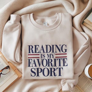 Może przedstawiać: Beżowa bluza z napisem "READING IS MY FAVORITE SPORT" w kolorze granatowym i czerwonym. Bluza jest złożona na miękkim, kremowym kocu, z książką i okularami w tle.