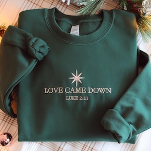 Puede incluir: Sudadera verde oscuro con el texto bordado "LOVE CAME DOWN" y "LUKE 2:11" en color claro. Un diseño de estrella está bordado encima del texto. La sudadera está doblada, con las mangas y los puños visibles.