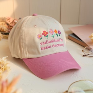 May include: A baseball cap with a cream-coloured crown and a pink peak. The cap has embroidered flowers in pink, orange, and purple, with the text "radicalized by basic decency" in pink.