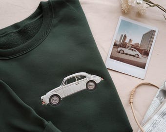 May include: Dark green sweatshirt with an embroidered white vintage car. A polaroid photo of a similar car in a city setting is next to the sweatshirt. The sweatshirt has a crew neck and long sleeves.