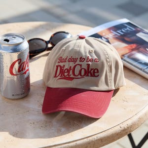 Może przedstawiać: Beżowa czapka z daszkiem z czerwonym daszkiem i haftem "Bad day to be a Diet Coke". Obok puszka Diet Coke i okulary przeciwsłoneczne.