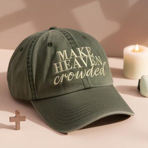 May include: Olive green baseball cap with the embroidered text "MAKE HEAVEN crowded" in white. The cap has a curved brim and a classic six-panel design. A lit candle and a small wooden cross are in the background.