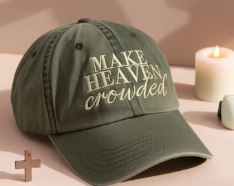May include: Olive green baseball cap with the embroidered text "MAKE HEAVEN crowded" in white. The cap has a curved brim and a classic six-panel design. A lit candle and a small wooden cross are in the background.