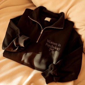 May include: Black quarter-zip sweatshirt with a gold-coloured zip. The text "Women in Psychology" is embroidered in a light colour on the front, with a small bow graphic. The sweatshirt is folded on a beige surface.