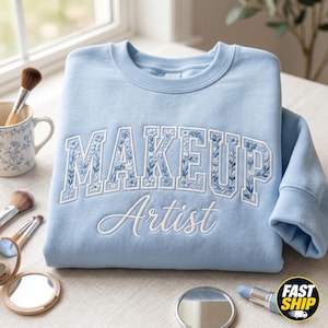 Pode incluir: Um moletom azul claro com as palavras "MAKEUP Artist" bordadas em branco. A palavra "MAKEUP" tem um design floral. Pincéis de maquiagem, um espelho compacto e batom são dispostos ao redor do moletom.