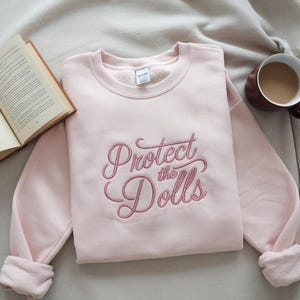 Könnte beinhalten: Ein rosa Sweatshirt mit dem Text "Protect the Dolls", der in rosa auf der Vorderseite gestickt ist.