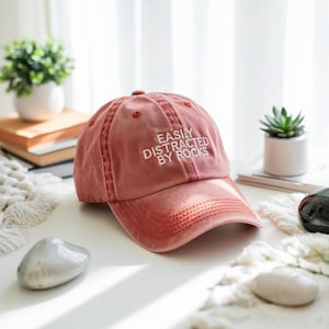 May include: A dusty rose baseball cap with the text "EASILY DISTRACTED BY ROCKS" embroidered in white. The cap is sitting on a white surface with decorative rocks and small potted plants in the background.
