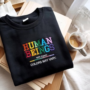 May include: A black sweatshirt with a rainbow embroidered design that reads "HUMAN BEINGS 100% Organic COLORS MAY VARY."