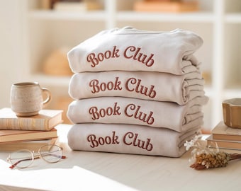 May include: A stack of four beige sweatshirts embroidered with the words "Book Club" in a reddish-brown script. The sweatshirts are neatly folded and stacked on a light-colored wooden surface, with books and a mug in the background.