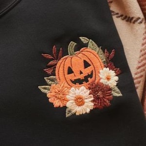 Peut inclure: Sweat-shirt noir orn&eacute; d'une broderie de citrouille souriante, entour&eacute;e de fleurs et de feuilles aux couleurs automnales. Le motif comprend des &eacute;l&eacute;ments floraux orange, blancs et bordeaux, cr&eacute;ant un th&egrave;me d'automne.