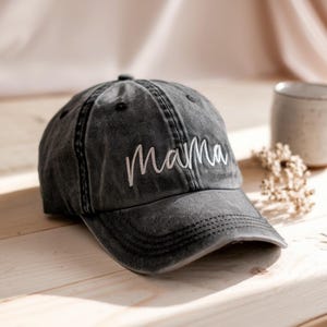 Può includere: Un berretto da baseball nero consumato con testo ricamato bianco che dice "mamma".