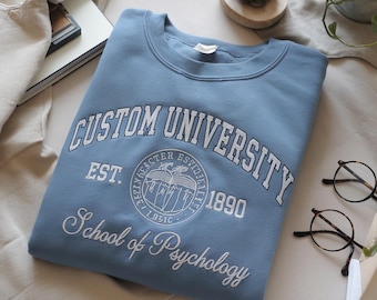 May include: A light blue sweatshirt with the words "CUSTOM UNIVERSITY" embroidered in white. Below the text is a circular emblem and the words "School of Psychology". The sweatshirt is folded neatly on a white surface.