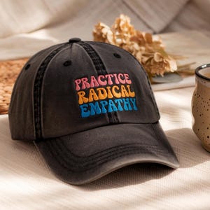 May include: A dark gray baseball cap with the embroidered text "PRACTICE RADICAL EMPATHY" in pink, orange, and blue. The cap has a curved brim and a classic six-panel design. The hat is sitting on a light-colored surface.