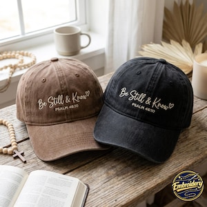 Könnte beinhalten: Zwei Baseballkappen, eine braune und eine schwarze, jeweils mit dem gestickten weißen Text "Be Still & Know", gefolgt von "PSALM 46:10" und einem kleinen Herzsymbol. Beide Kappen haben eine strukturierte Krone, einen gebogenen Schirm und einen Knopf.
