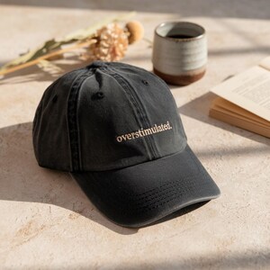 May include: A black baseball cap with the text "overstimulated." embroidered in a light brown color.